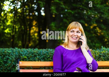 Defocus bionda caucasica sorridente donna che parla al telefono all'esterno, all'aperto. donna di 40 anni in blusa viola in panchina. fe. Adulto Foto Stock
