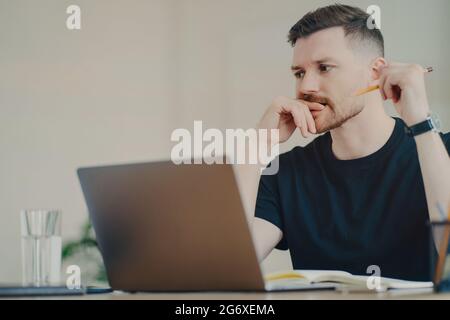 L'uomo serio allievo bearded ama studiare il processo ha i lavori di espressione ponderati freelance vestiti in camicia nera casuale di t scrive giù gli usi delle note Foto Stock