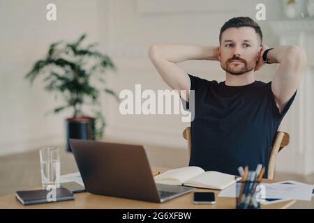 Il freelance maschile pensivo prende la pausa tiene le mani dietro le pose testa ai lavori desktop sul computer portatile ha espressione pensierosa pensa al futuro Foto Stock