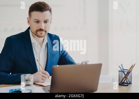 Il marketer serio dell'uomo bearded coinvolto nel processo di lavoro fa le note con la matita osserva attentamente al calcolatore del laptop indossa il vestito formale blu pone a. Foto Stock