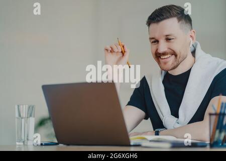 L'uomo felice bearded prepara il lavoro di corso di carta funziona a distanza da casa orologi webinar utilizza connessione Internet ad alta velocità computer portatile e auricolari moderni Foto Stock