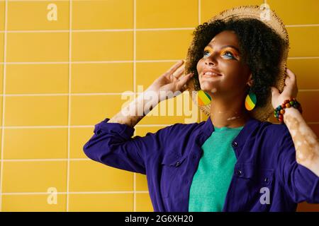 Donna sorridente che indossa un cappello di paglia e vestiti alla moda guardando in piedi vicino al muro giallo dell'edificio Foto Stock