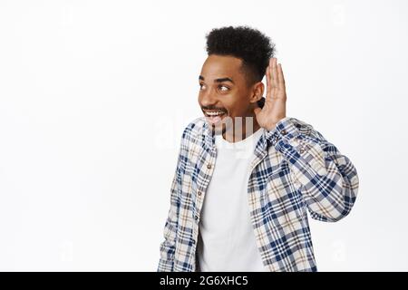 Sorridi un ragazzo afro-americano maledetto, che si avvicina con la mano vicino all'orecchio e grinning, sorprendi interessanti pettegolezzi, in piedi su bianco Foto Stock