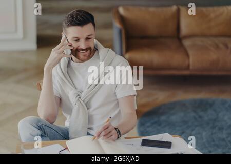 Ritratto di uomo sorridente in t-shirt bianca coperta di maglione, seduto sulla sedia e parlando al telefono Foto Stock