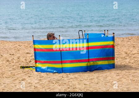 Bournemouth, Dorset UK. 9 luglio 2021. Tempo nel Regno Unito: Bella giornata calda e soleggiata, ma umida alle spiagge di Bournemouth, mentre i cercatori di sole si dirigano verso il mare per godersi il sole. Uomo dietro colorato vento frangivento a righe break vento sulla sabbia sulla spiaggia. Credit: Carolyn Jenkins/Alamy Live News Foto Stock