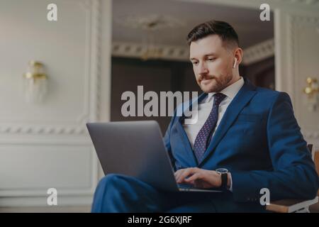 Uomo d'affari serio in tuta blu che indossa auricolari wireless seduti in soggiorno con il computer in ginocchio Foto Stock