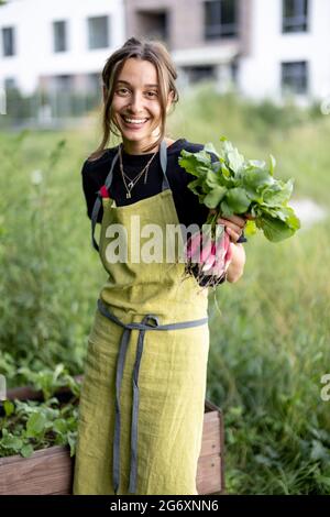 Donna felice che tiene il ravanello fresco nelle mani e che mostra alla macchina fotografica vicino al giardino domestico. Cibo biologico sano, verdure, agricoltura, primo piano. Foto Stock