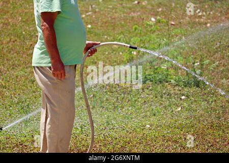 L'uomo con un tubo in mano innaffia l'erba. Il giardiniere si prende cura del prato in tempo caldo Foto Stock