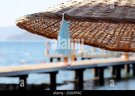 Maschere facciali protettive appese sul parasolo di vimini sul mare e sullo sfondo del molo. Sicurezza su una spiaggia durante la pandemia del coronavirus, vacanza estiva Foto Stock