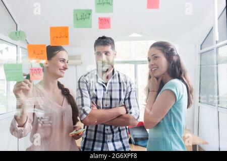 Visione a basso angolo di giovani lavoratori affidabili che inviano promemoria sul muro trasparente di un ufficio, accanto al loro collega in un moderno spazio di lavoro Foto Stock