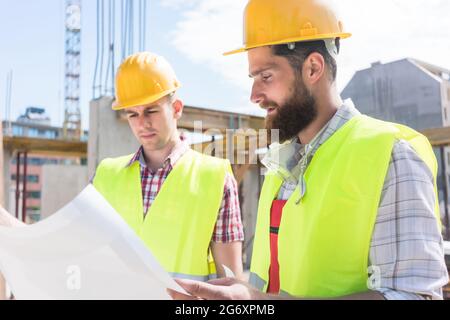 Due giovani operai edili che indossano cappelli rigidi gialli e giubbotti di sicurezza riflettenti mentre analizzano insieme il progetto di un nuovo edificio Foto Stock