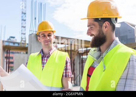 Due giovani operai edili che indossano cappelli rigidi gialli e giubbotti di sicurezza riflettenti mentre analizzano insieme il progetto di un nuovo edificio Foto Stock