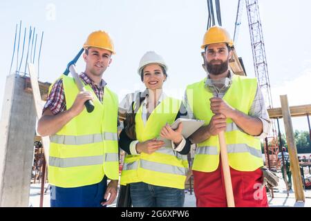 Ritratto di tre colleghi fiduciosi che si pongono insieme indossando cappelli di sicurezza duri e giubbotti riflettenti durante il lavoro sul cantiere di una c Foto Stock