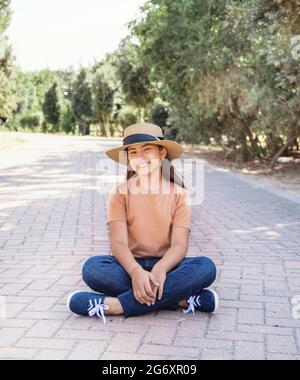 Un ragazzino sorridente che indossa t-shirt, jeans e cappello di paglia siede all'aperto nel parco Foto Stock