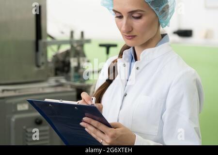 Ritratto di un ispettore femminile che scrive osservazioni sull'attrezzatura e sul processo di produzione durante il controllo di qualità in una fabbrica di cosmetici Foto Stock