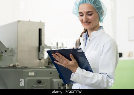 Ritratto di un ispettore femminile che scrive osservazioni sull'attrezzatura e sul processo di produzione durante il controllo di qualità in una fabbrica di cosmetici Foto Stock
