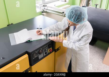 Vista laterale di un ingegnere femminile che scrive osservazioni per un rapporto sulle attrezzature e la produzione, mentre si trova vicino a una macchina di compressore in un moderno Foto Stock
