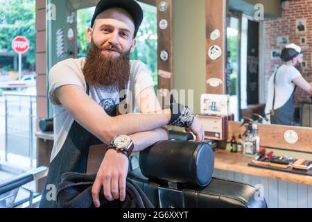 Ritratto di un fiducioso giovane barbiere sorridente e guardando la telecamera, mentre appoggiata su un hair styling in cattedra all'interno di un vintage salone di bellezza Foto Stock