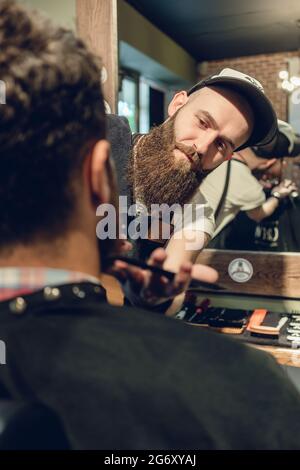 Vista laterale ravvicinata della testa di un giovane e delle mani di un esperto acconciatore che regola la barba, con pettine e forbici in un salone di bellezza Foto Stock