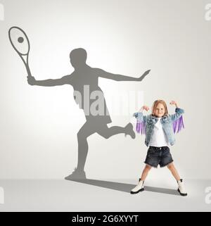 Infanzia e sogno sul grande e famoso futuro. Immagine concettuale con la ragazza e l'ombra del tennista femminile su sfondo grigio chiaro. Foto Stock