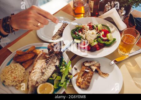 L'uomo mangia pesce con verdure e insalata greca nel ristorante all'aperto. Cena a base di pesce fresco servita. Primo piano vista dall'alto del cibo Foto Stock