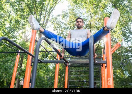Vista ad angolo basso di un bel giovane atletico che esercita la gamba verticale solleva su barre parallele in un parco fitness all'aperto con attrezzature moderne Foto Stock