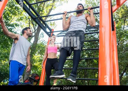 Vista ad angolo basso di un giovane forte che fa ripetizioni chin-up per la parte superiore del corpo durante l'allenamento calistenico, con i suoi amici in un moderno fi esterno Foto Stock