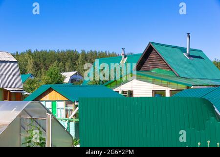 Tetti di case in legno e serre nel villaggio sullo sfondo del cielo blu e verde fogliame. Caldo sole giorno estivo. Piano medio Foto Stock