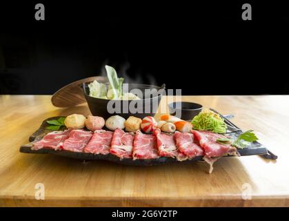 Set di carni fresche grezze di manzo o di maiale shabu e condimento con zuppa calda in ciotola nera su tavolo di legno Foto Stock