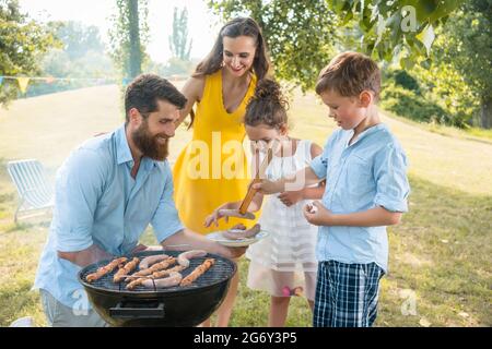 Padre dedicato che aiuta suo figlio ad usare le pinze di legno per mettere le salsicce sulla griglia barbecue al carbone all'aperto durante il picnic di famiglia in estate Foto Stock