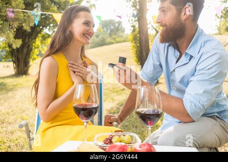 Giovane uomo bello seduto sul ginocchio mentre mostra la sua bella ragazza un anello di fidanzamento romantico durante un picnic con vino rosso e fresco Foto Stock