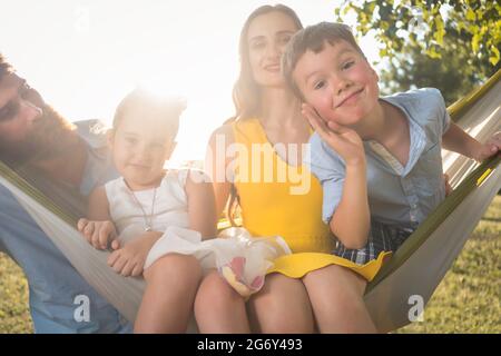 Carino bambino che fa un volto divertente, mentre guardando la fotocamera per un ritratto di famiglia con la sorella e i genitori su un amaca all'aperto in una giornata di sole summ Foto Stock