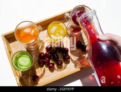 Un vassoio di legno con bevande colorate: Limonata arancione, gialla e cetriolo. Nelle vicinanze si trovano le ciliegie. Una mano versa il succo rosso da un decanter in un bicchiere. C Foto Stock