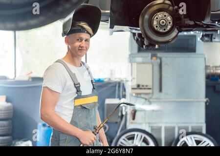 Ritratto di un giovane e bel meccanico auto che guarda la fotocamera con sicurezza durante la saldatura durante il lavoro in un moderno negozio di riparazione auto Foto Stock