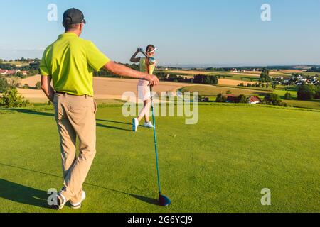 Vista posteriore completa di un uomo che indossa abiti da golf moderni, mentre il suo partner colpisce la palla durante la partita sull'erba verde di un professionista Foto Stock