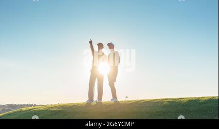 A basso angolo di visione della silhouette di un uomo rivolto verso l'orizzonte, mentre in piedi accanto al suo partner femmina su un campo di golf professionale contro il sole Foto Stock