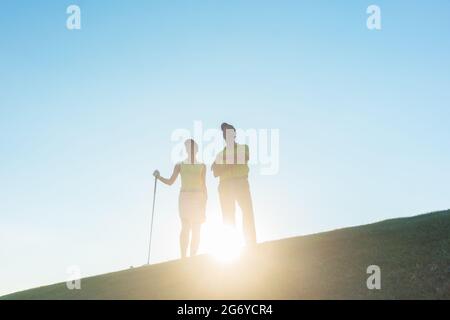 Vista ad angolo basso della silhouette di un uomo che punta all'orizzonte mentre si trova accanto alla sua partner femminile, su un campo da golf professionale contro il sole Foto Stock