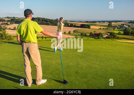 Vista posteriore completa di un uomo che indossa abiti da golf moderni mentre guarda il suo compagno colpire la palla, durante la partita sull'erba verde di un professionista Foto Stock