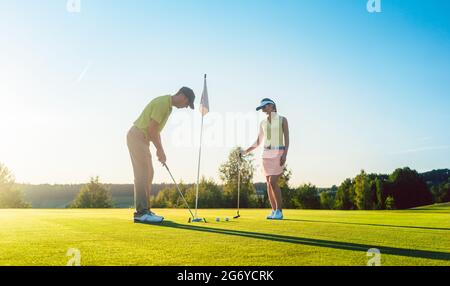 Vista laterale completa di un uomo pronto a colpire la palla da golf nel buco, mentre si esercita il tiro corto con il suo compagno di gioco in un paese moderno clu Foto Stock