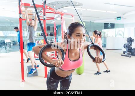 Giovane donna allegra che si allena con anelli ginnici durante l'allenamento di routine in un centro fitness alla moda con attrezzature moderne per traini funzionali Foto Stock