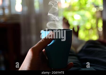 Tazza blu di caffè in mano dell'uomo a letto a casa. Uomo sdraiato e rilassante in una calda giornata estiva. Sfondo verde offuscato della porta aperta e vista sul giardino Foto Stock