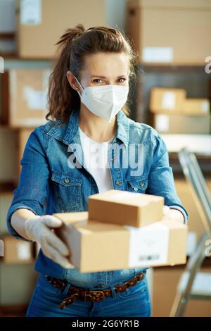 Consegna commerciale. Donna moderna in jeans con maschera ffp2 che dà pacchi in magazzino. Foto Stock