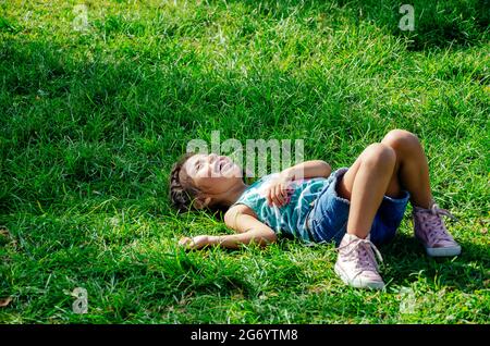 Una bambina sorride sdraiata sull'erba verde brillante. Ritratto di un bambino felice sull'erba in estate. La vista dall'alto. Spazio per il testo. Foto Stock