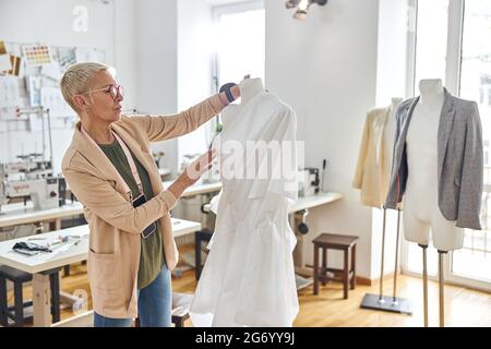 Meditorosa dressmaker matura guarda il tessuto bianco con pinned raccoglie su manichino in officina Foto Stock