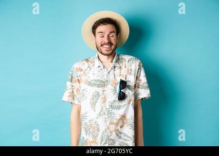 Felice turista godendo di vacanza estiva, con cappello di paglia e camicia hawaiana, in piedi gioioso su sfondo blu Foto Stock