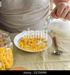 L'uomo corto versa il latte in cornflakes in un piatto bianco su una tovaglia liscia e ruvida. Vista dall'alto. Messa a fuoco selettiva. Concetto, semplice veloce americano h Foto Stock