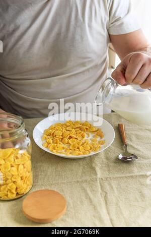 L'uomo corto versa il latte in cornflakes in un piatto bianco su una tovaglia liscia e ruvida. Vista dall'alto. Messa a fuoco selettiva. Concetto, semplice veloce americano h Foto Stock