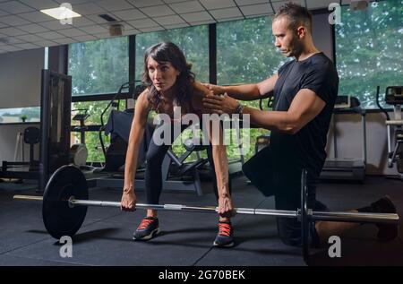 Primo piano ritratto di forte e concentrata donna fitness giovane in sportswear che fa il sollevamento del peso di potenza in palestra interna mentre personal trainer Foto Stock