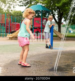 I bambini sorridenti che giocano con l'acqua al paraspruzzi nel parco pubblico locale nelle calde giornate estive. Piccoli bei bambini che si divertono al fo Foto Stock