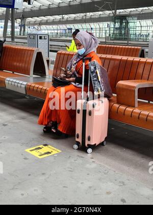 Amsterdam, Paesi Bassi, 25 giugno 2021 : Signora in un abito marocchino arancione con una sciarpa e una maschera alla bocca, seduta accanto ai suoi bagagli su una panchina nella sala della stazione di Amsterdam Bijlmer Arena. Sul terreno poco prima di lei il cartello della distanza di 1,5 m. Foto Stock